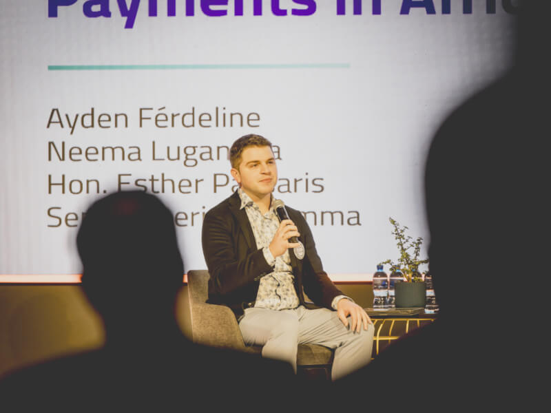 Ayden Férdeline holding a microphone, seated on a chair on stage, giving a presentation. Behind him, part of his slide is visible on a large screen. In the foreground, the silhouettes of audience members are visible between the camera and the speaker.