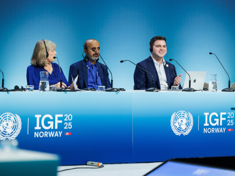 Three participants seated at tables with microphones, engaged in discussion during the Internet Governance Forum; Ayden Ferdeline is among them.